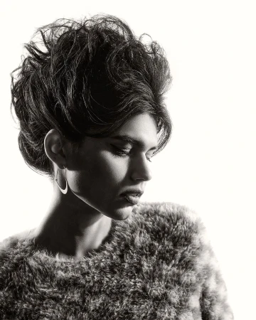 Monochrome portrait of a person with dark hair in an updo wearing hoop earrings and a textured fur-like top against a white backdrop.